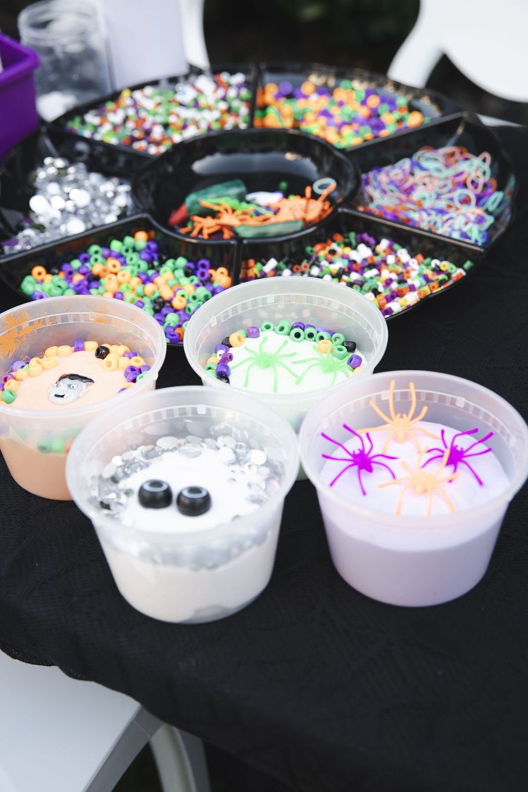 Four finished slime cups decorated with beads, googly eyes and toy spiders next to a tray of colorful toppings